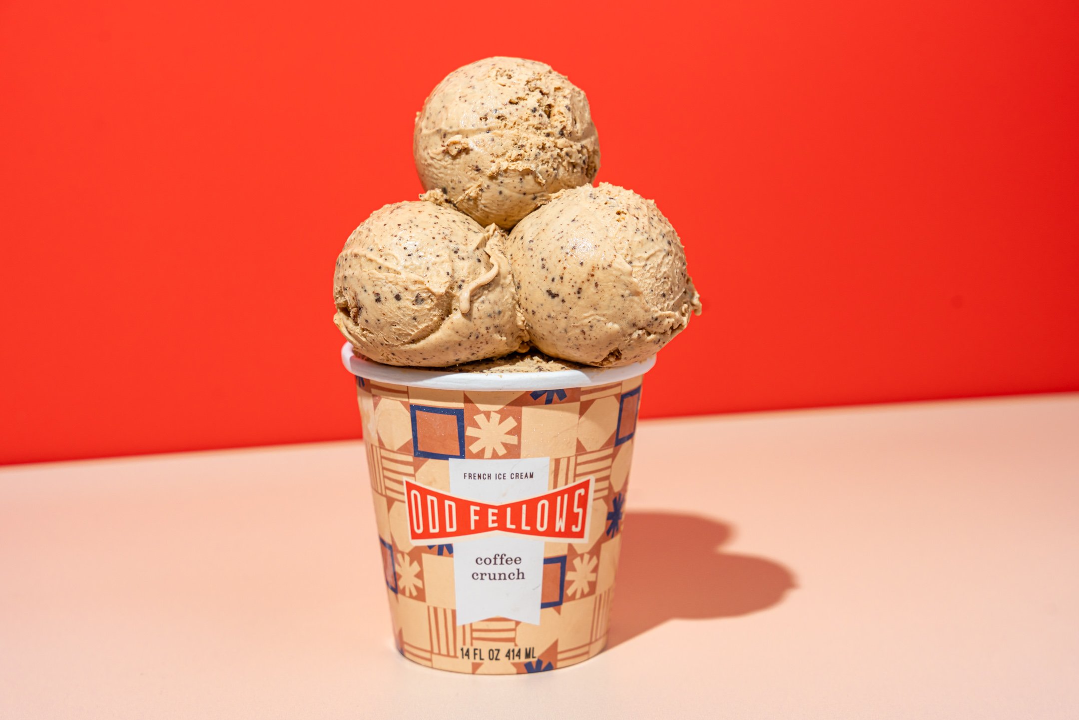 A cup of Odd Fellows coffee ice cream with three scoops on top, placed on a light pink surface against a red background.