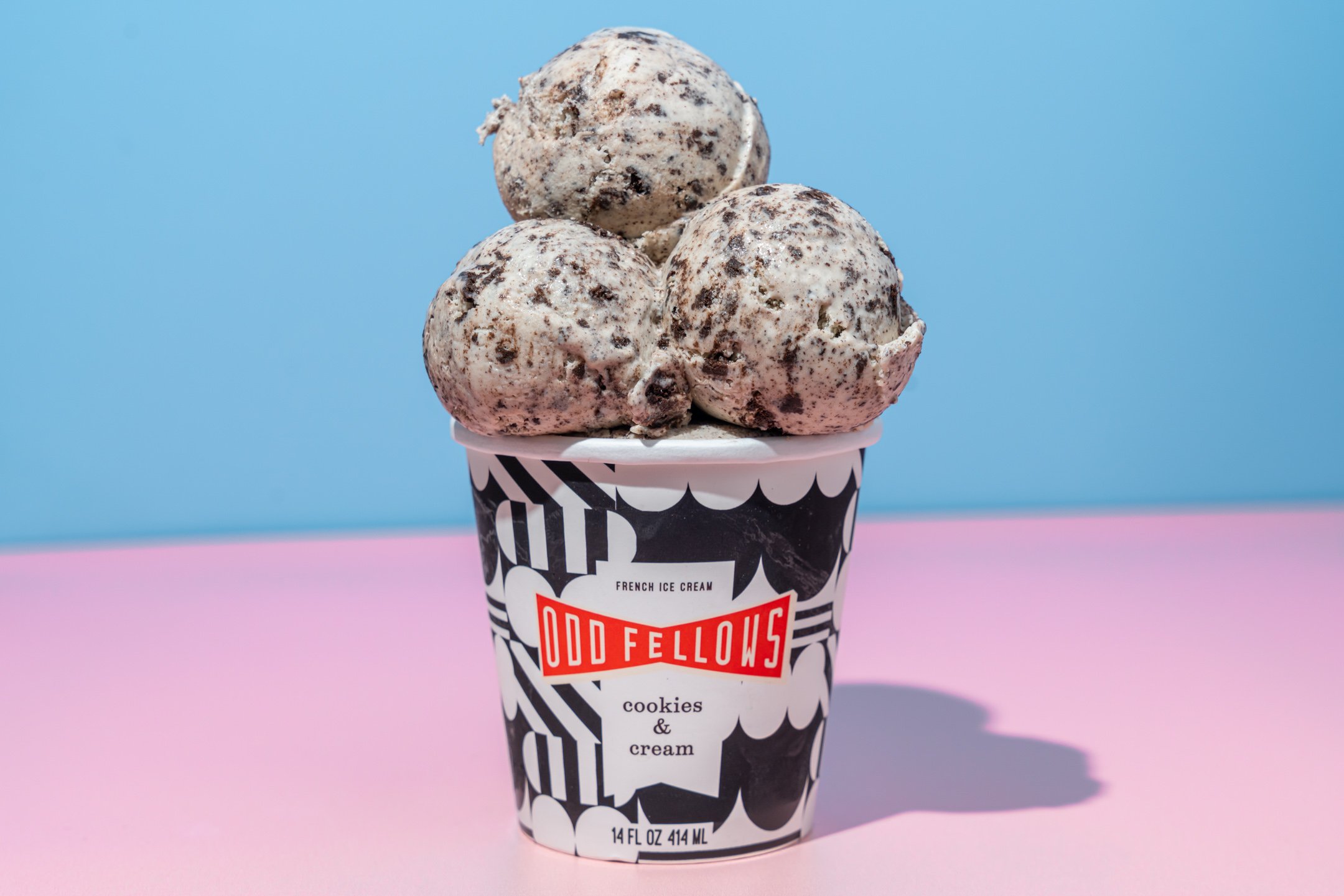 A cup of Odd Fellow's cookies and cream ice cream topped with three scoops of cookies and cream ice cream with cookie chunks, against a pink and blue background.