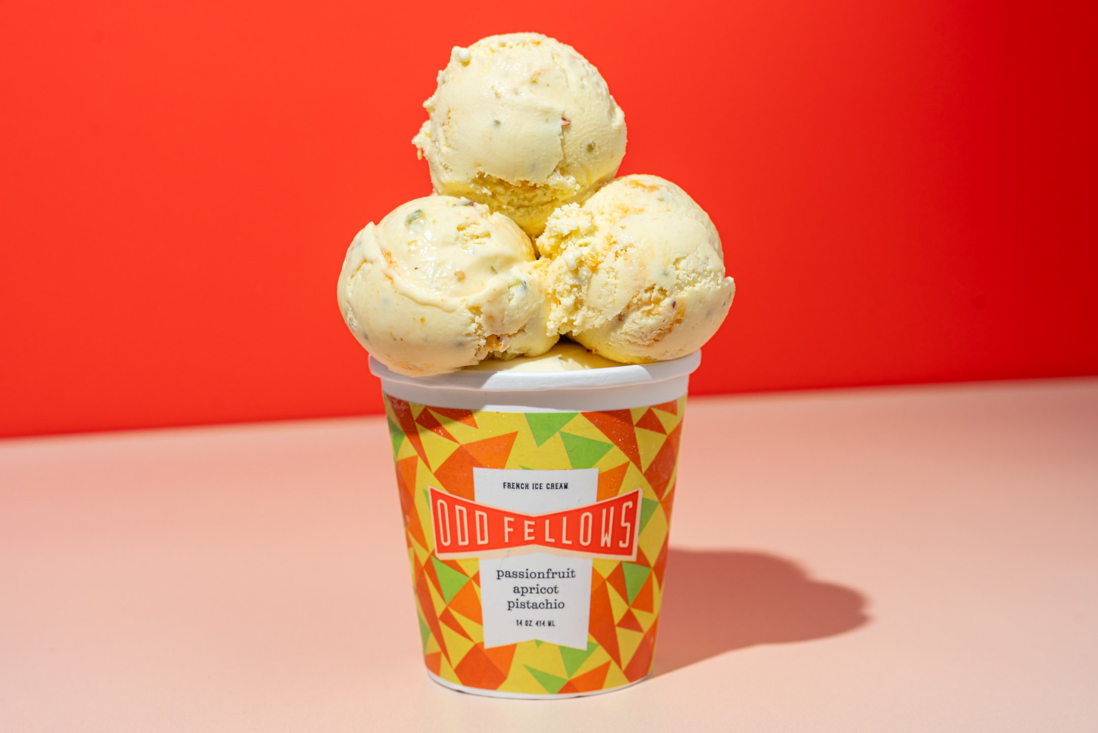 Three scoops of passionfruit, apricot, and pistachio ice cream in a colorful cup against a red and pink background.