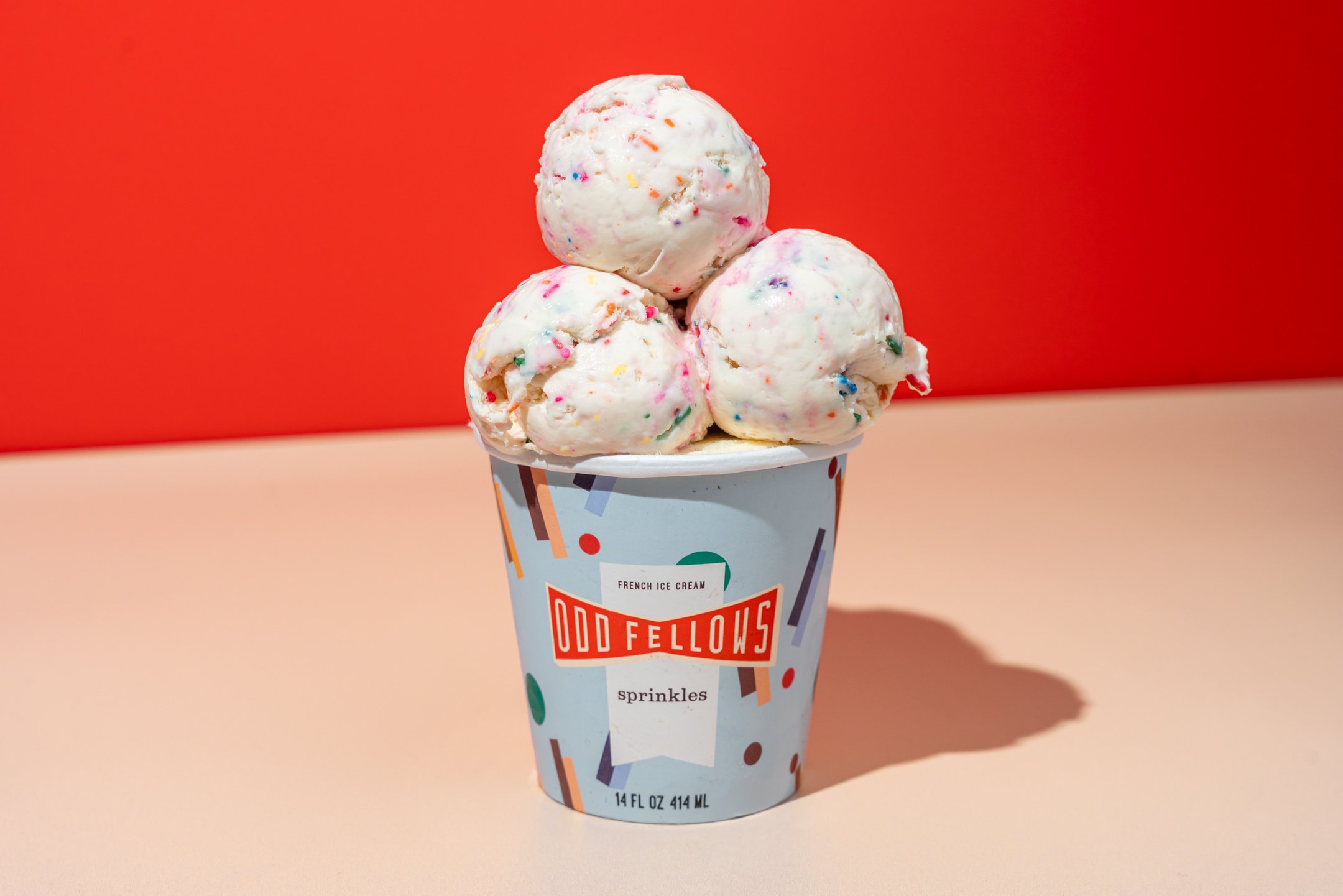 A cup of vanilla ice cream with colorful sprinkles in an Odd Fellows branded cup, against a red and pink background.