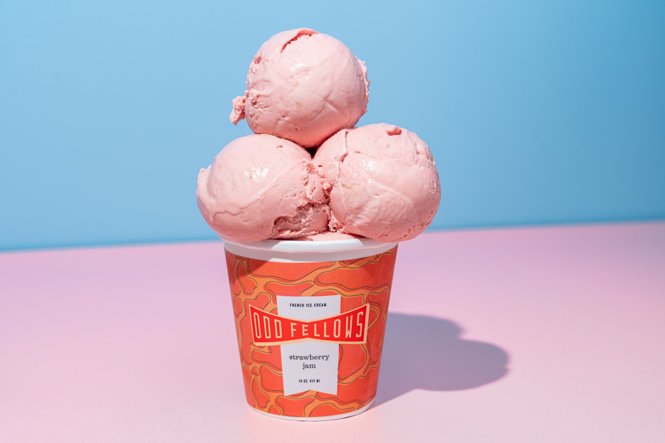 Three scoops of pink strawberry ice cream in a branded cup on a pink surface with a blue background.